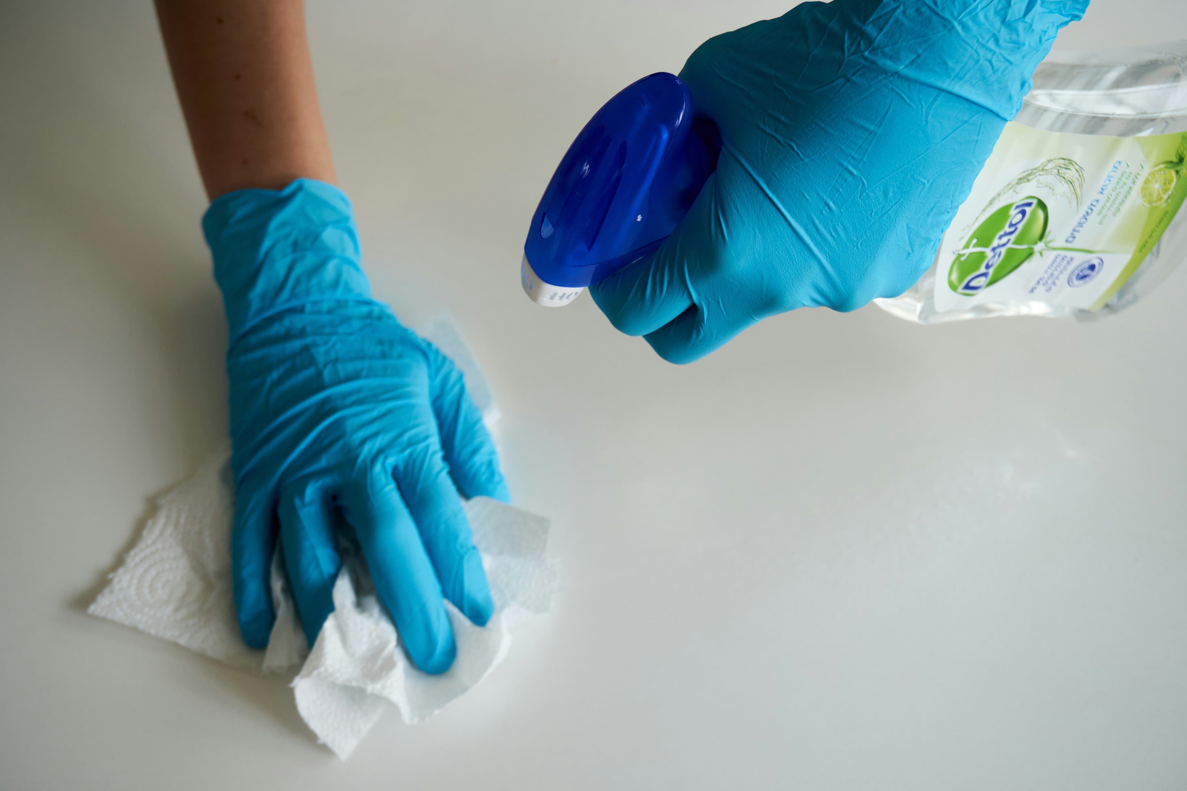 Two gloved hands clean a white surface with a spray bottle and a white cloth.