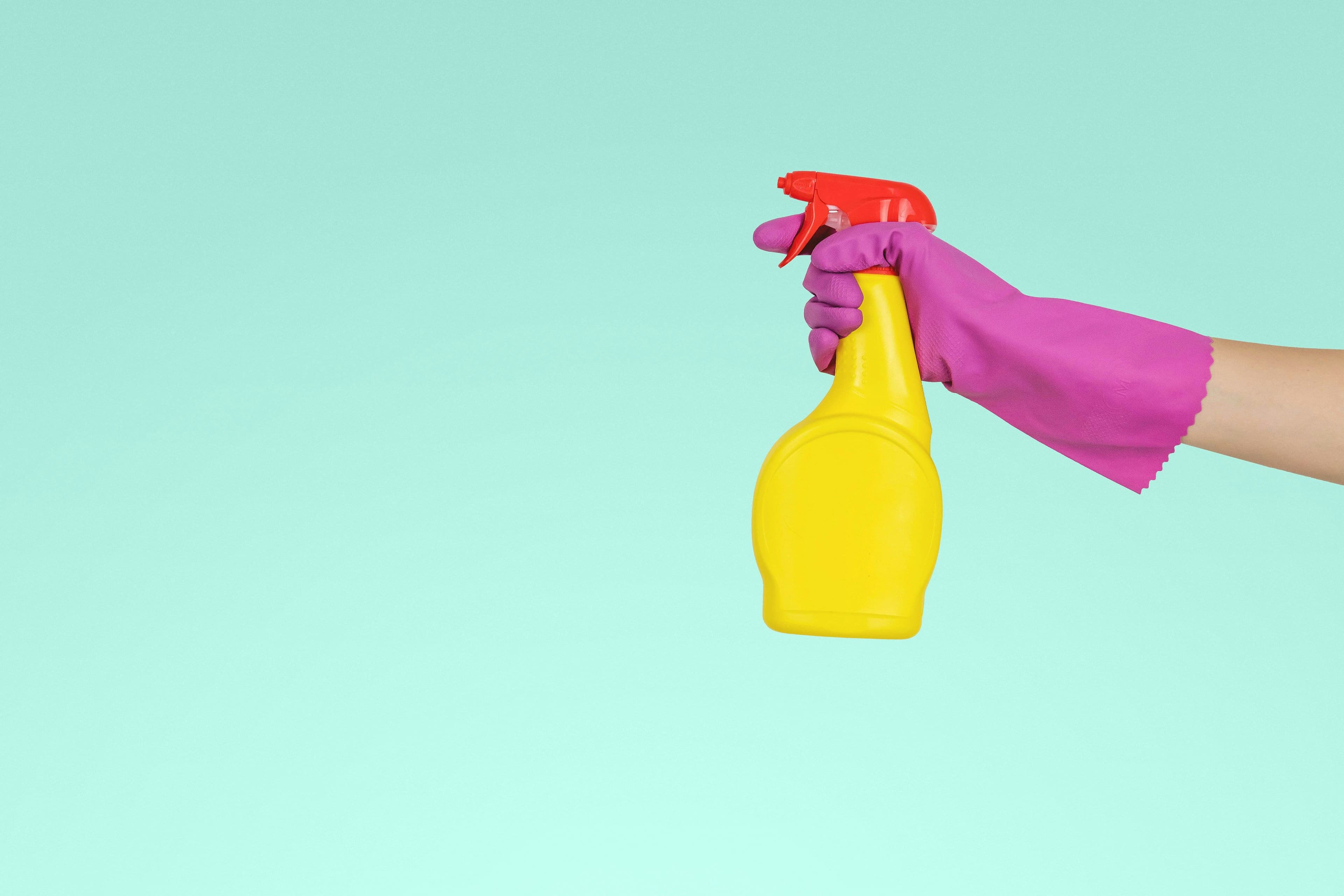 Hand in pink rubber glove holding a yellow spray bottle against a light blue background.